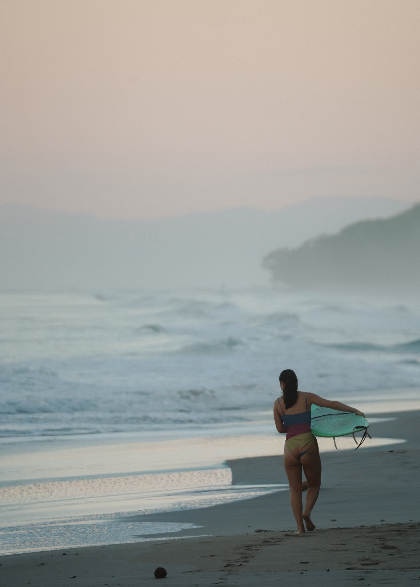 A solo surfer walking along the beach at sunrise with her board